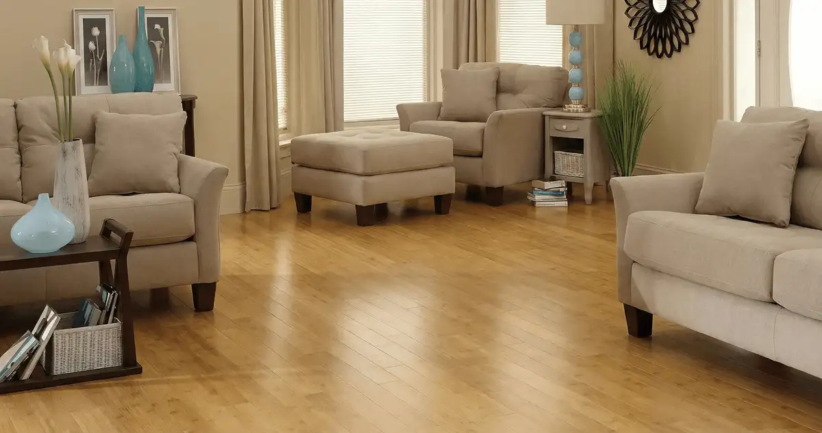 A living room with professionally installed bamboo flooring, beige sofas, a matching ottoman, a side table with a blue lamp, vases, framed pictures, and large windows with blinds and curtains in Jersey City, NJ